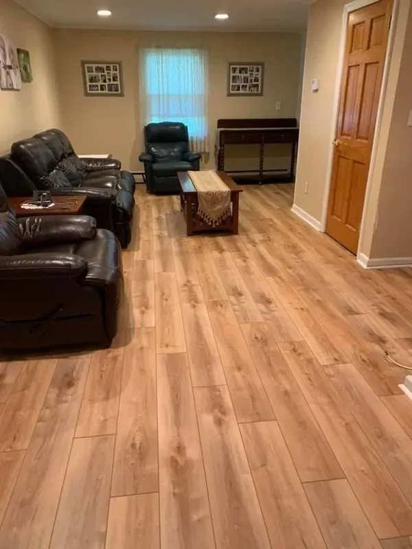 A living room with light brown wood laminate flooring, dark brown leather sofas, a dark green recliner, and a wooden coffee table with a fringed runner, and a brown wooden door in Livingston, NJ